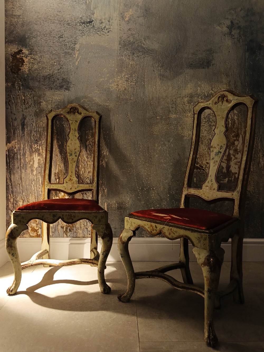 A pair of 18th-century North Italian side chairs featuring distressed cream painted frames with carved crests, cabriole legs, and red velvet seats.