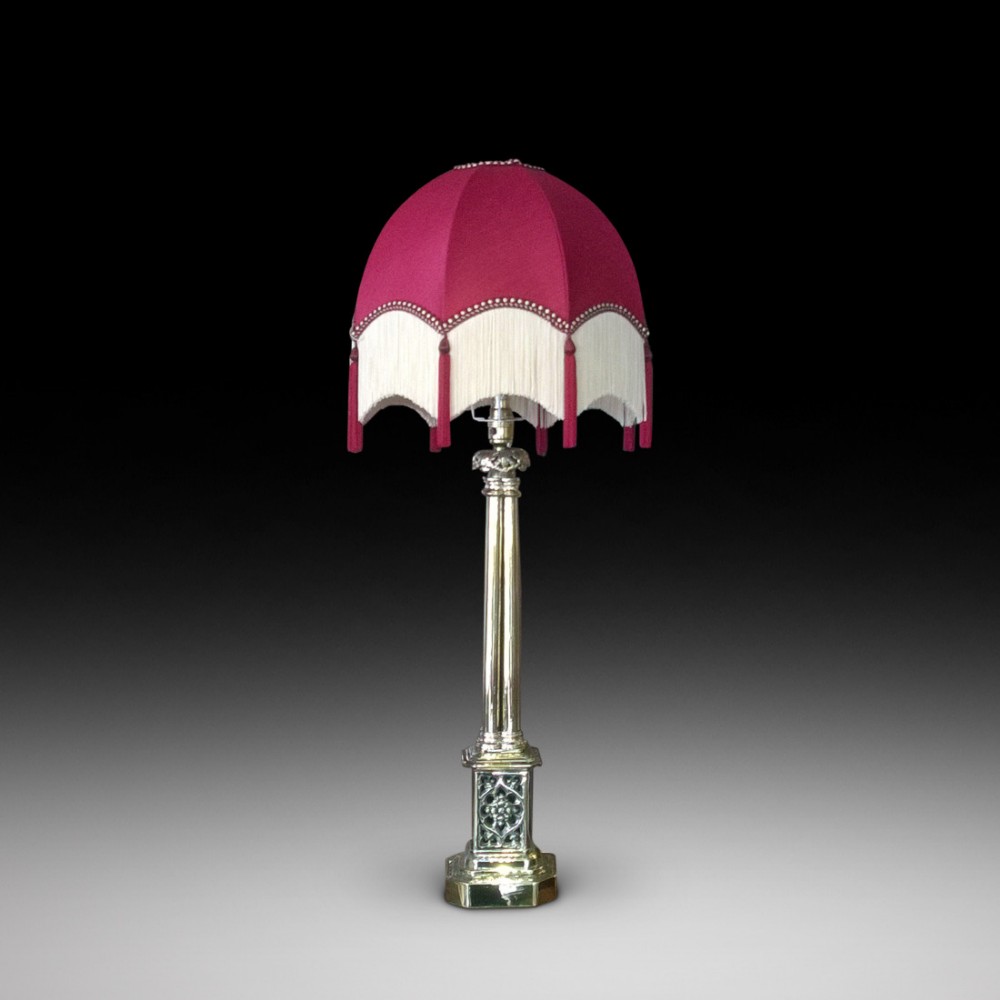 An Edwardian polished brass column table lamp with a decorative plinth base and a magenta fabric shade with cream fringe.