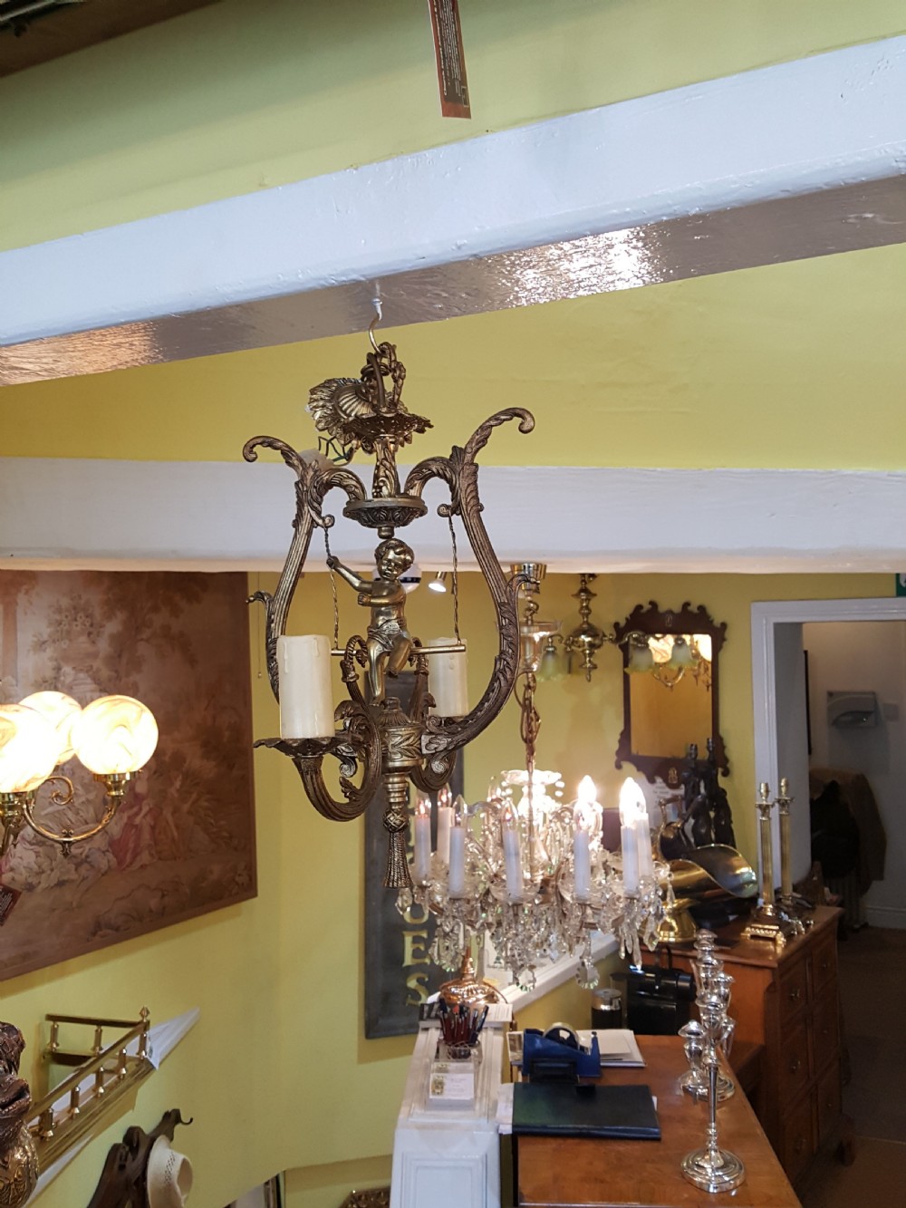 A pair of gilt metal ceiling chandeliers featuring a central figure of a cherub seated on a swing within an ornate lyre-shaped frame with two candle arms. View II