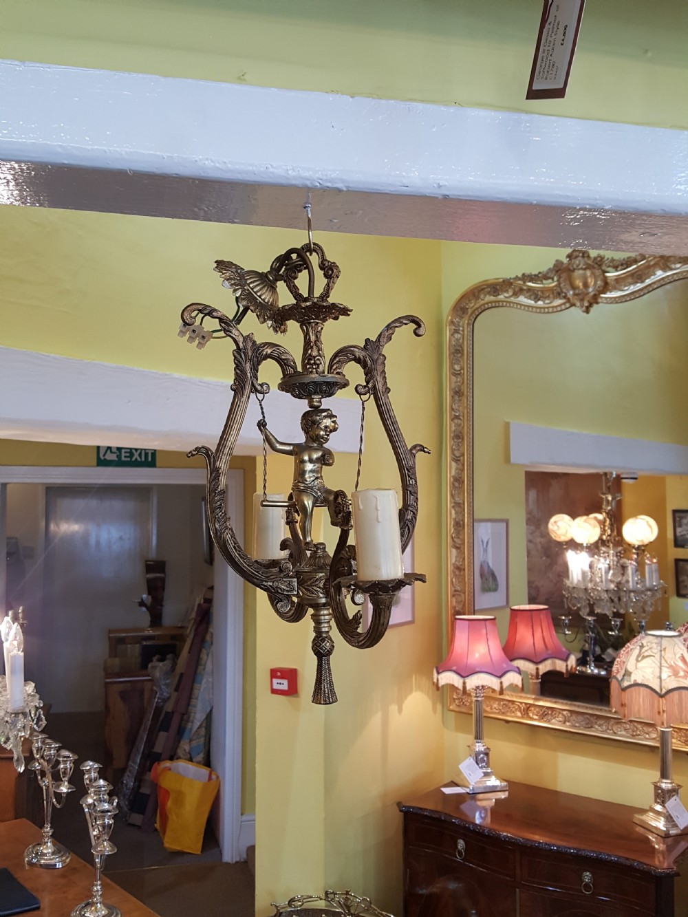 A pair of gilt metal ceiling chandeliers featuring a central figure of a cherub seated on a swing within an ornate lyre-shaped frame with two candle arms. View III