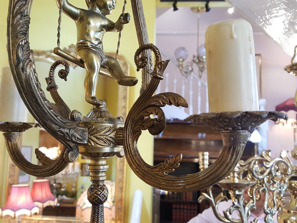 A pair of gilt metal ceiling chandeliers featuring a central figure of a cherub seated on a swing within an ornate lyre-shaped frame with two candle arms. View IX