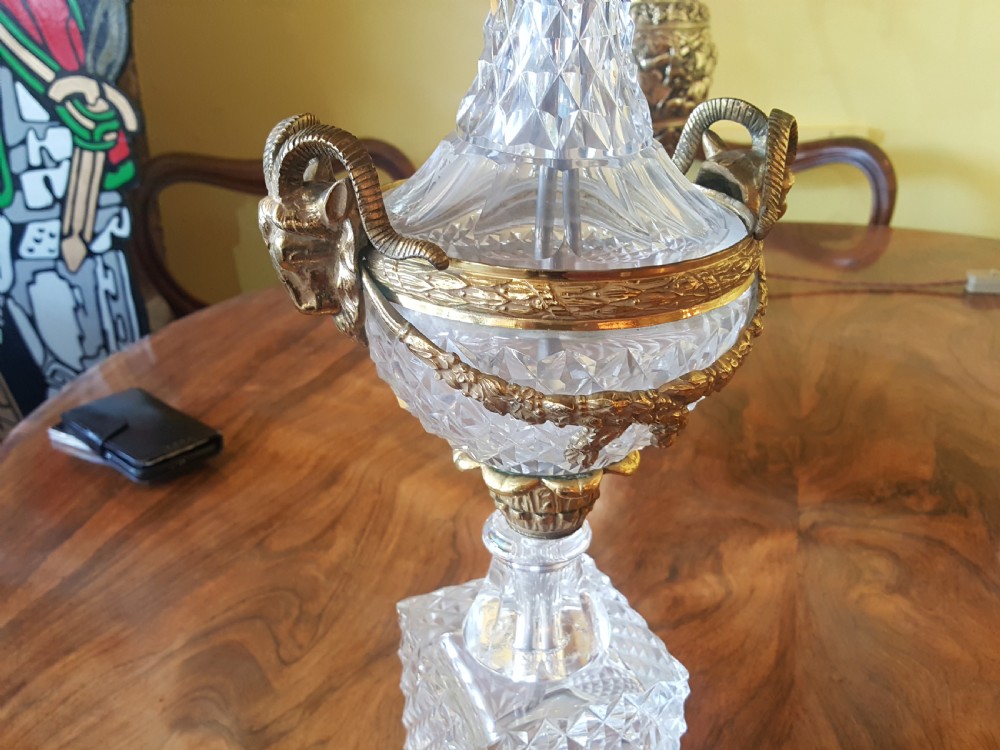 A cut glass urn-form table lamp with ornate gilt metal handles and base, topped with a black and gold patterned domed shade featuring long black fringe trim. View V