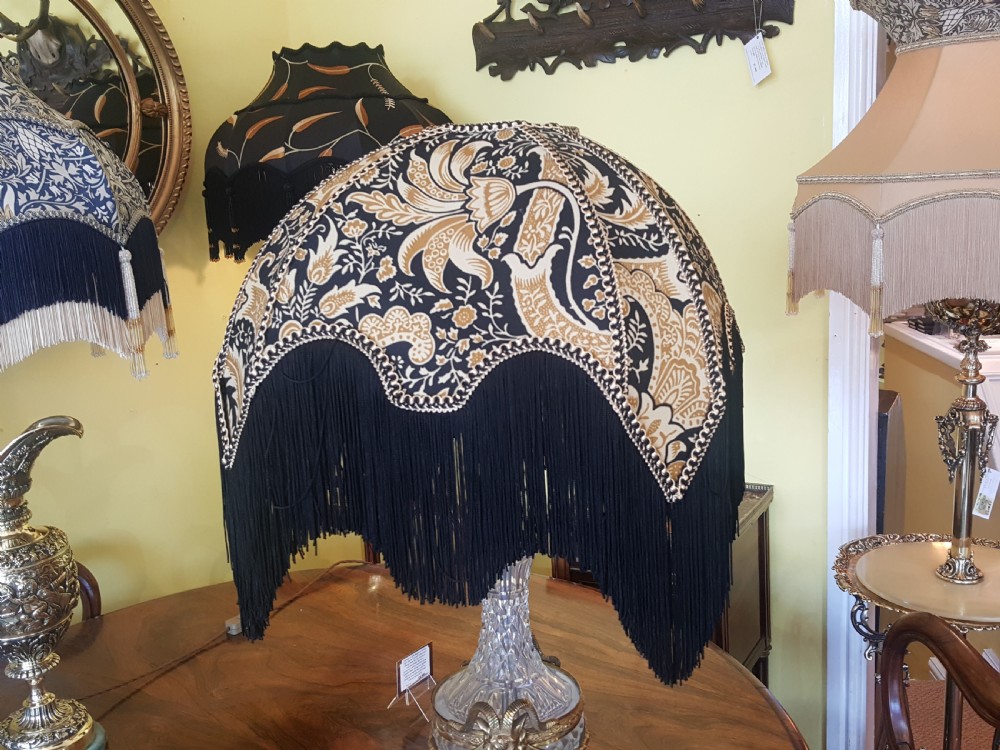 A cut glass urn-form table lamp with ornate gilt metal handles and base, topped with a black and gold patterned domed shade featuring long black fringe trim. View VI