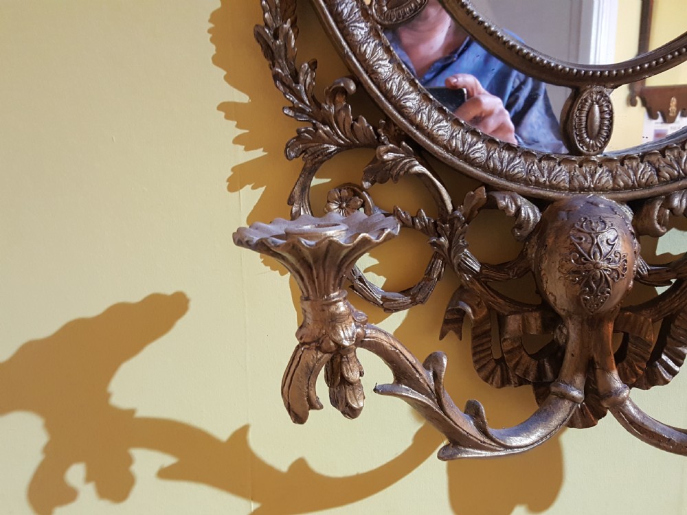 An ornate oval giltwood girandole mirror featuring carved acanthus cresting, ribbon detailing, and two candle sconces at the base. View IV