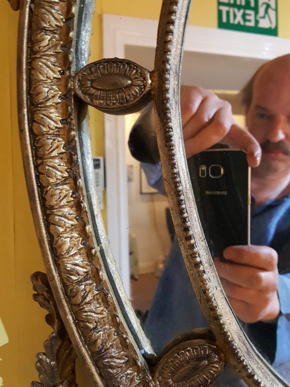 An ornate oval giltwood girandole mirror featuring carved acanthus cresting, ribbon detailing, and two candle sconces at the base. View V