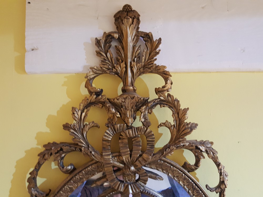 An ornate oval giltwood girandole mirror featuring carved acanthus cresting, ribbon detailing, and two candle sconces at the base. View VI