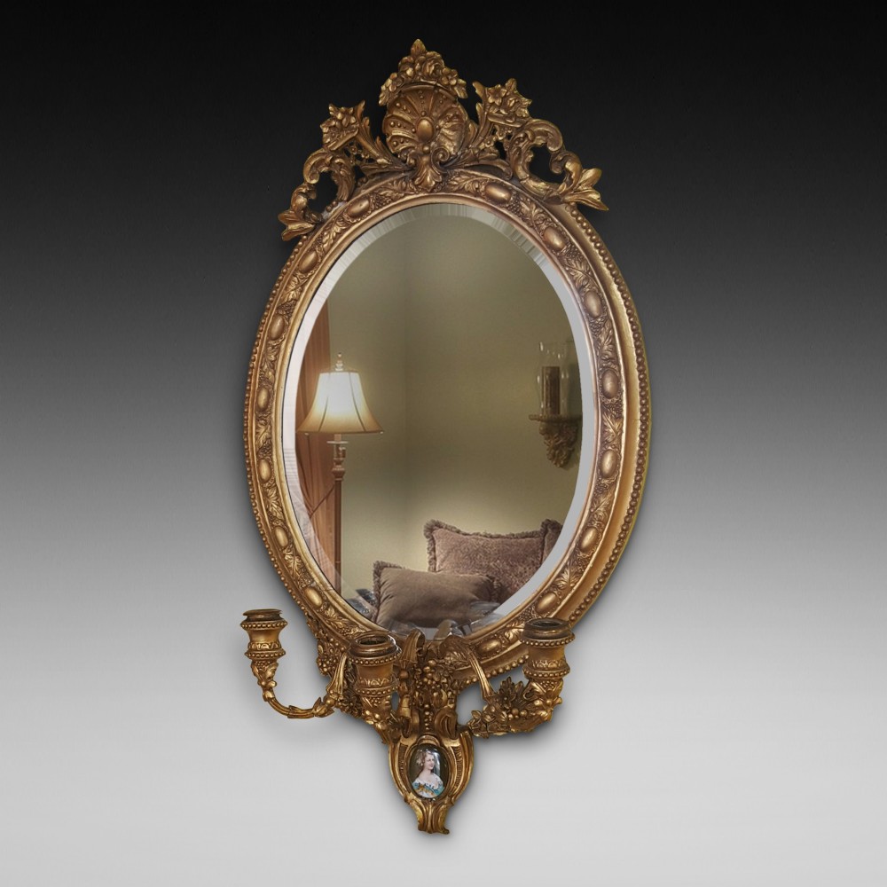 A 19th-century oval giltwood wall mirror featuring an ornate shell cresting, candle sconces at the base, and an inset miniature portrait.