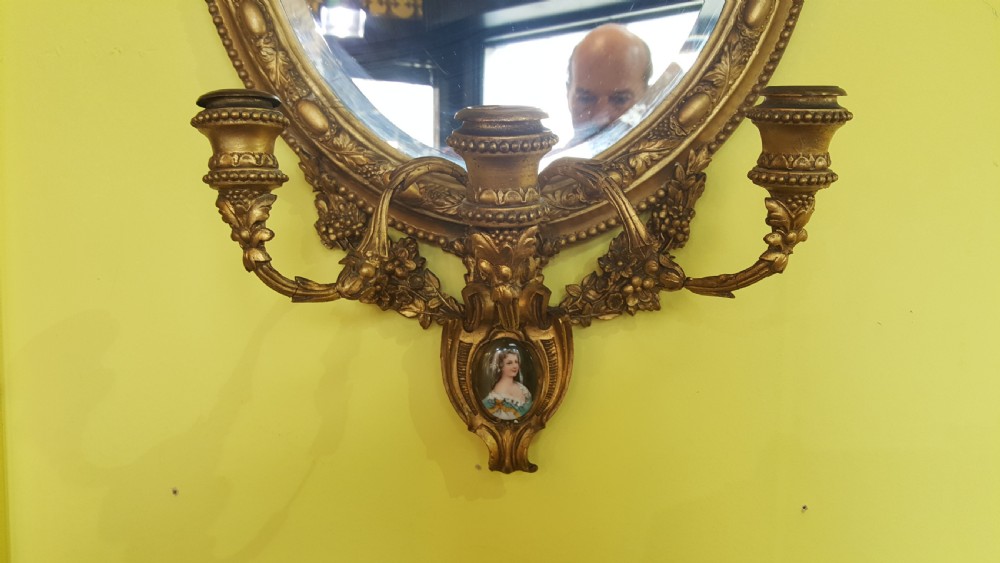 A 19th-century oval giltwood wall mirror featuring an ornate shell cresting, candle sconces at the base, and an inset miniature portrait. View III