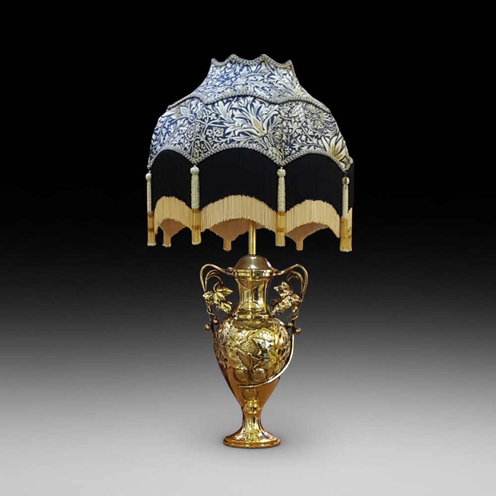 An Art Nouveau brass table lamp shaped like an urn with foliate handles and an intricate patterned fabric shade with decorative fringe.