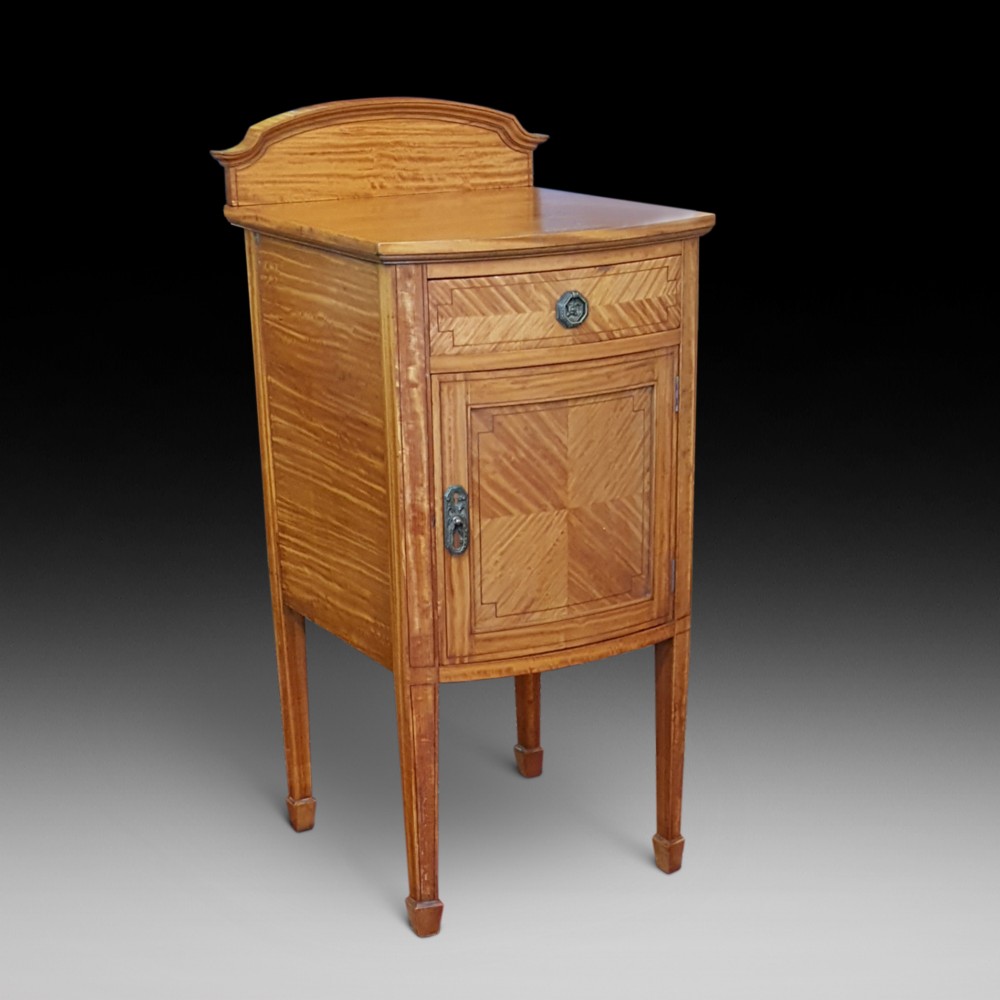 An antique Sheraton Revival satinwood bow-fronted bedside cabinet featuring a top drawer above a single cupboard door with geometric parquetry on square tapered legs.