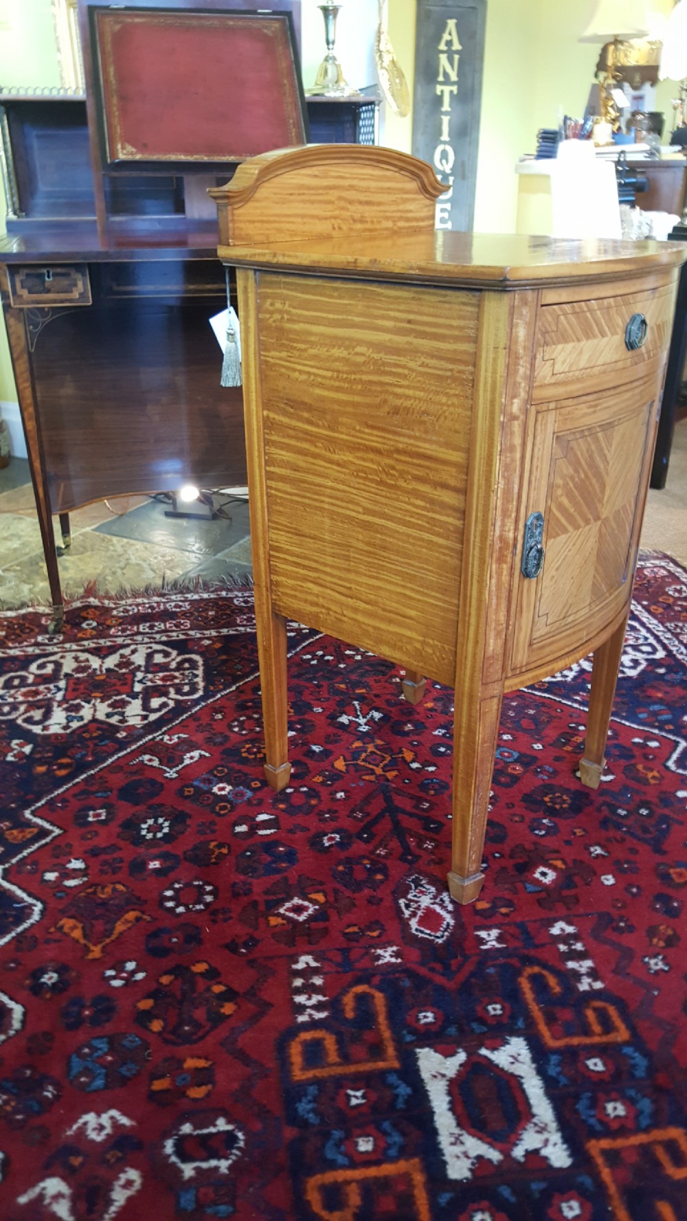 An antique Sheraton Revival satinwood bow-fronted bedside cabinet featuring a top drawer above a single cupboard door with geometric parquetry on square tapered legs. View II
