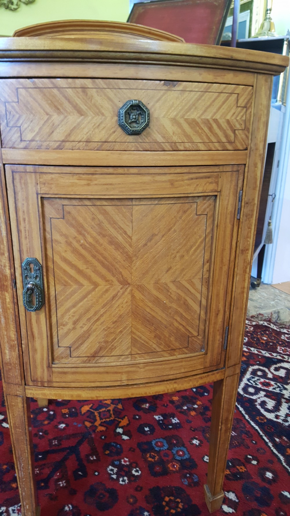 An antique Sheraton Revival satinwood bow-fronted bedside cabinet featuring a top drawer above a single cupboard door with geometric parquetry on square tapered legs. View III