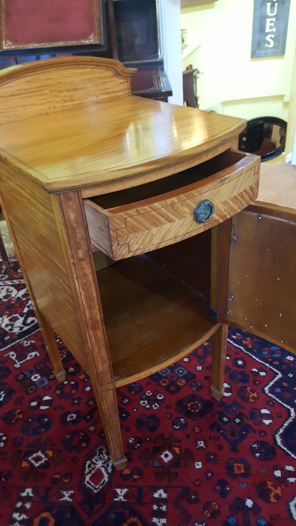 An antique Sheraton Revival satinwood bow-fronted bedside cabinet featuring a top drawer above a single cupboard door with geometric parquetry on square tapered legs. View VII