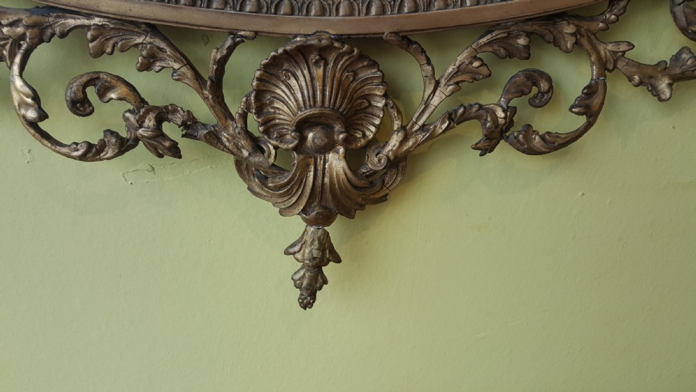 An oval Adam Revival giltwood mirror featuring a top crest with a classical urn, scrolling foliate detail, and a carved shell motif at the base. View II