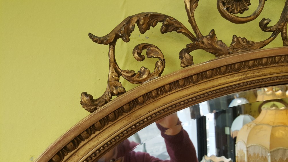 An oval Adam Revival giltwood mirror featuring a top crest with a classical urn, scrolling foliate detail, and a carved shell motif at the base. View IV