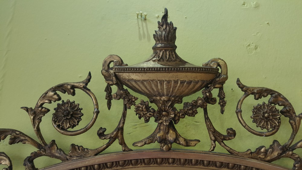 An oval Adam Revival giltwood mirror featuring a top crest with a classical urn, scrolling foliate detail, and a carved shell motif at the base. View V