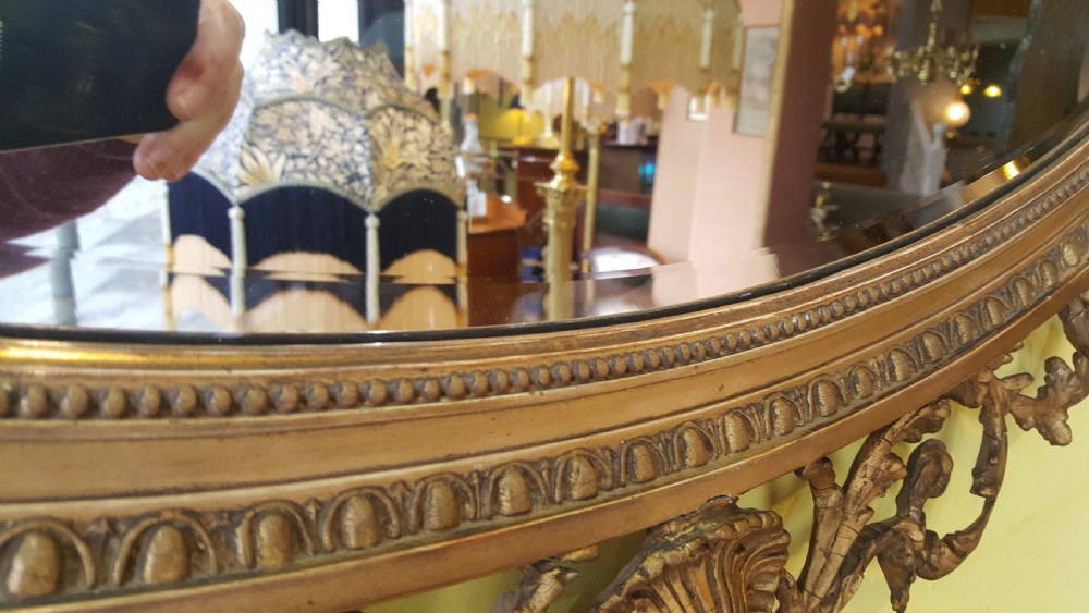 An oval Adam Revival giltwood mirror featuring a top crest with a classical urn, scrolling foliate detail, and a carved shell motif at the base. View VI