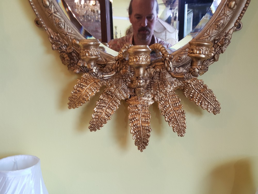 An oval Victorian giltwood wall mirror featuring prominent botanical leaf cresting and integrated candle sconces at the base. View II