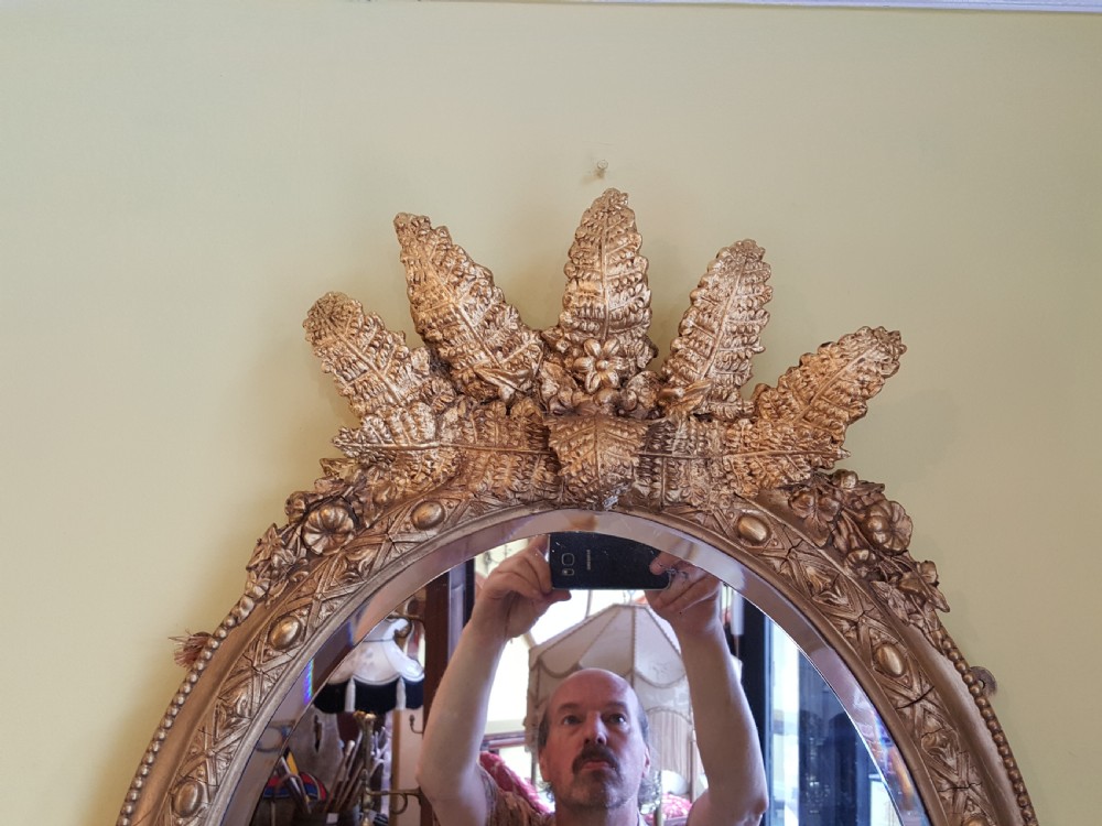 An oval Victorian giltwood wall mirror featuring prominent botanical leaf cresting and integrated candle sconces at the base. View III