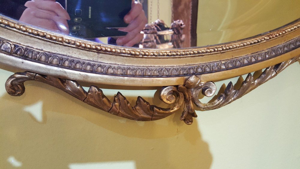An oval Adam Revival wall mirror featuring a gilt and gesso frame adorned with a ribbon knot cresting and foliate swags. View II