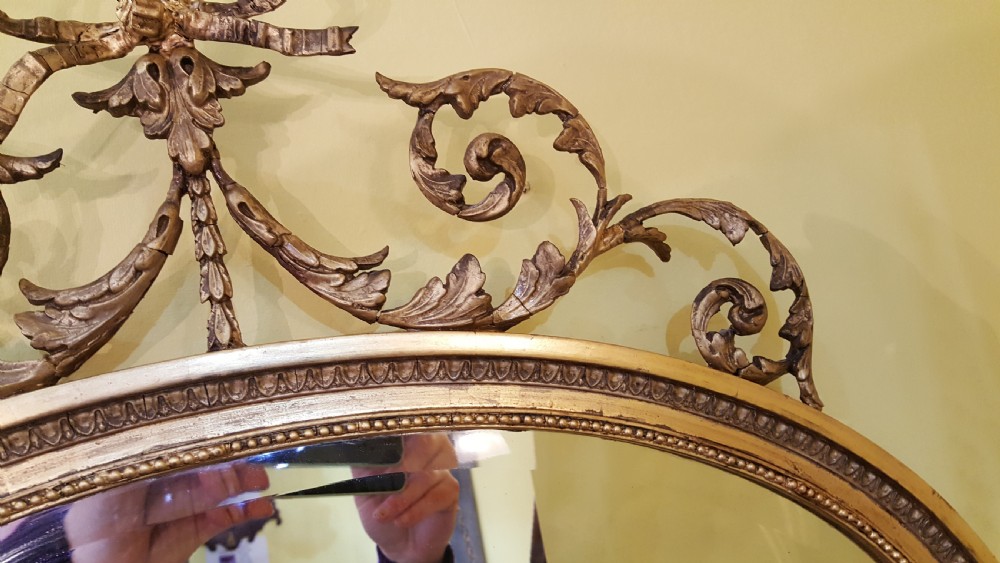 An oval Adam Revival wall mirror featuring a gilt and gesso frame adorned with a ribbon knot cresting and foliate swags. View V