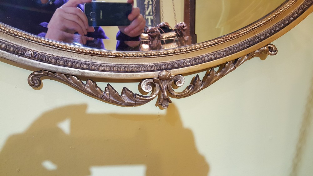 An oval Adam Revival wall mirror featuring a gilt and gesso frame adorned with a ribbon knot cresting and foliate swags. View VI