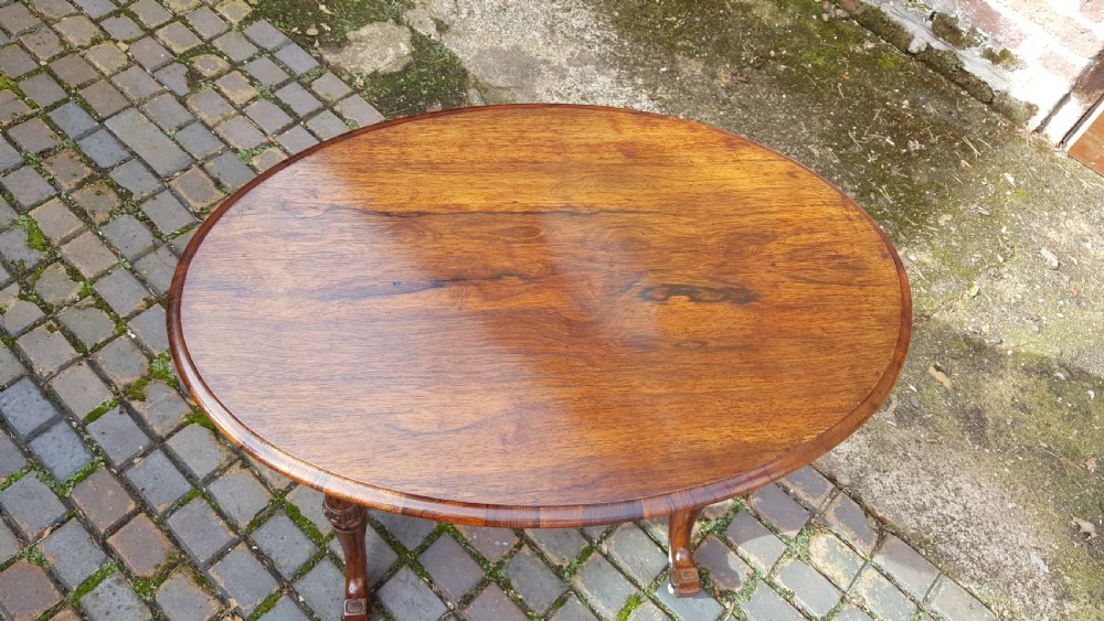 An oval Victorian rosewood occasional table featuring turned twin column legs, carved cross-stretchers, and small brass castors. View VI