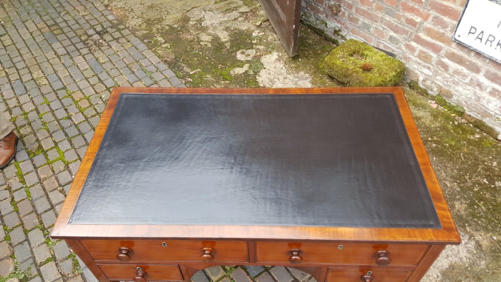 A George III mahogany writing desk featuring a black leather inset top, four drawers with turned wooden knobs, boxwood stringing inlay, an arched central apron, and square tapered legs on brass castors. View VI