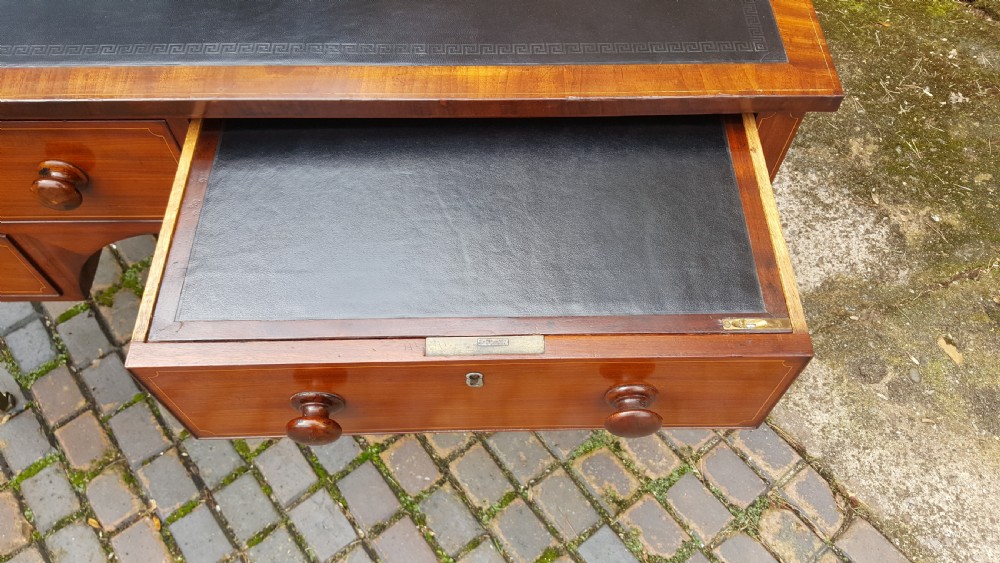 A George III mahogany writing desk featuring a black leather inset top, four drawers with turned wooden knobs, boxwood stringing inlay, an arched central apron, and square tapered legs on brass castors. View VII