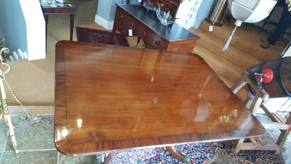 A Regency period rectangular mahogany tilt-top table with crossbanded edges supported by a central turned column and four reeded splayed legs on brass castors. View V