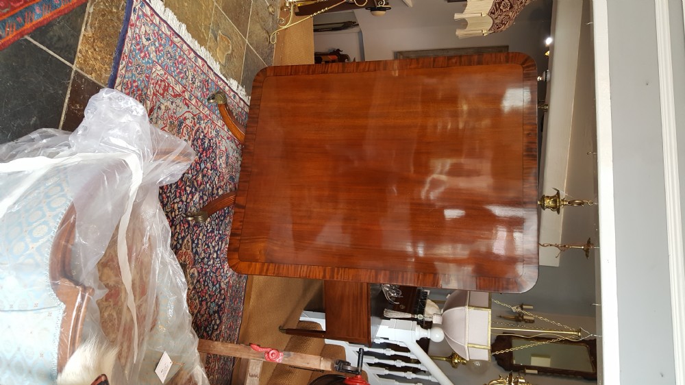 A Regency period rectangular mahogany tilt-top table with crossbanded edges supported by a central turned column and four reeded splayed legs on brass castors. View VI