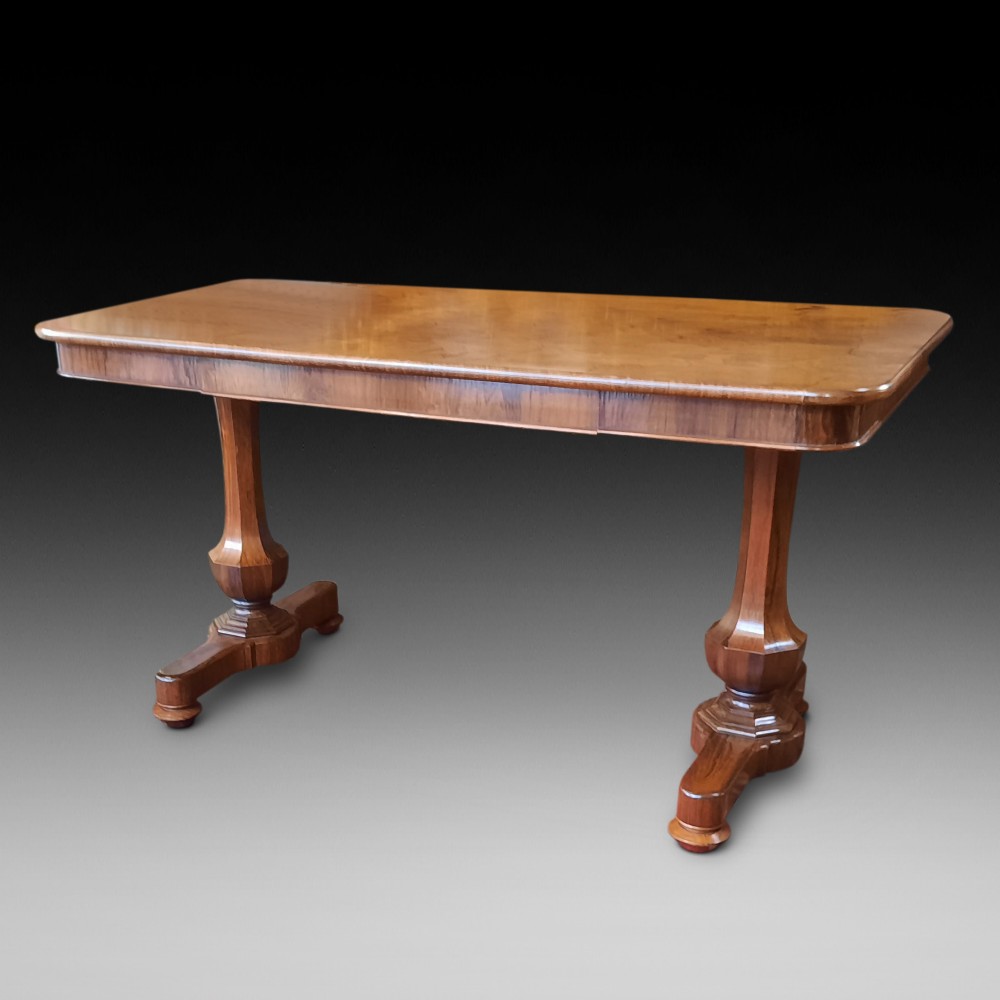 A Late Regency rosewood library table featuring a polished rectangular top with rounded corners, supported by dual faceted end supports resting on stepped plinth bases.