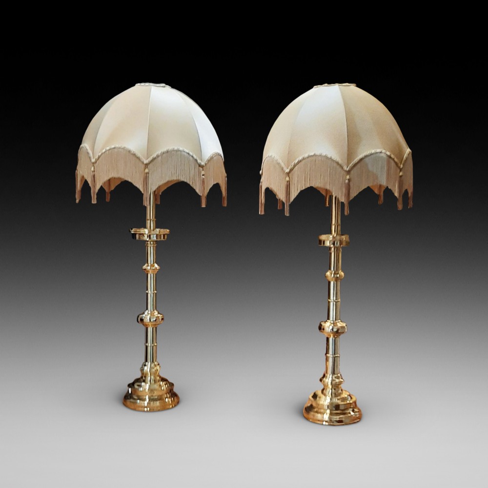 A pair of tall turned brass candlesticks fitted with dome-shaped beige fabric shades featuring scalloped fringe detail.