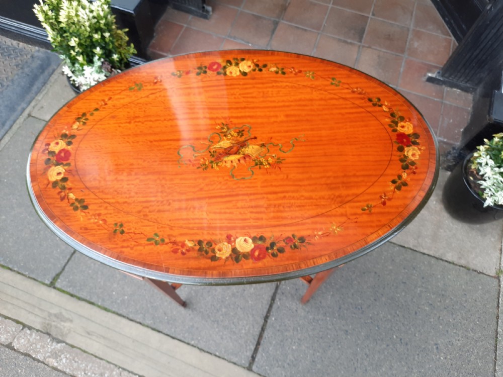 An Edwardian oval satinwood occasional table featuring hand-painted floral decoration on the top, slender tapered legs, and an X-form stretcher with a central wooden urn finial. View III