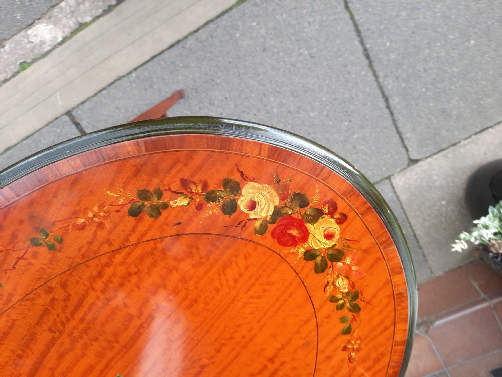 An Edwardian oval satinwood occasional table featuring hand-painted floral decoration on the top, slender tapered legs, and an X-form stretcher with a central wooden urn finial. View IV