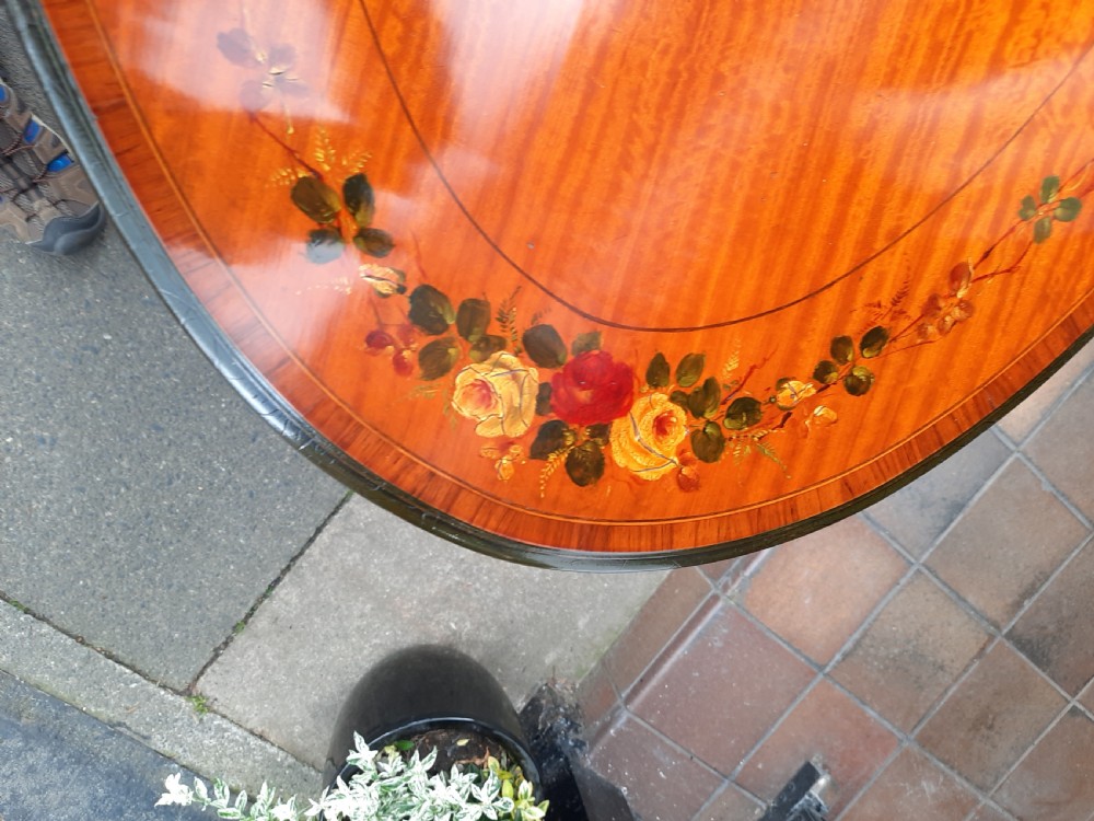 An Edwardian oval satinwood occasional table featuring hand-painted floral decoration on the top, slender tapered legs, and an X-form stretcher with a central wooden urn finial. View VII