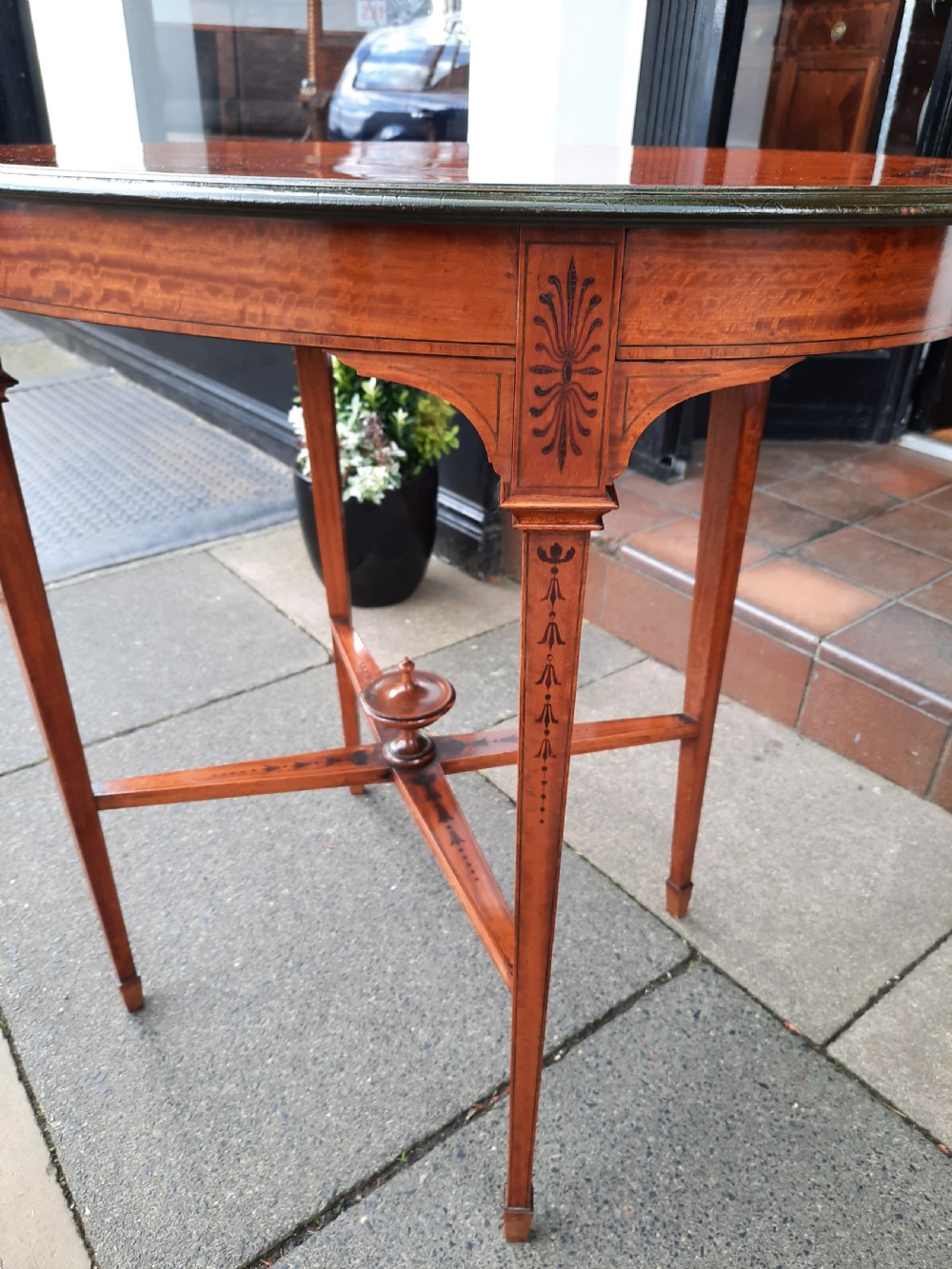 An Edwardian oval satinwood occasional table featuring hand-painted floral decoration on the top, slender tapered legs, and an X-form stretcher with a central wooden urn finial. View IX