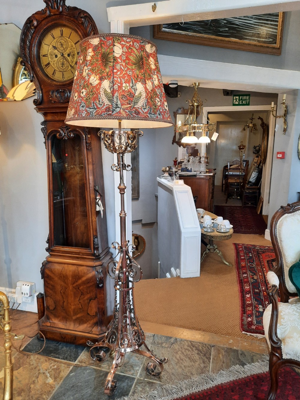 An Arts and Crafts copper floor lamp featuring an adjustable central stem, scrollwork base, and a patterned fabric shade. View VI