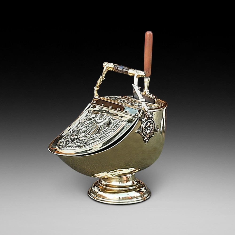 An antique brass coal purdonium in the Adam style, featuring intricate repousse classical motifs, a pedestal base, carrying handle, and attached shovel.
