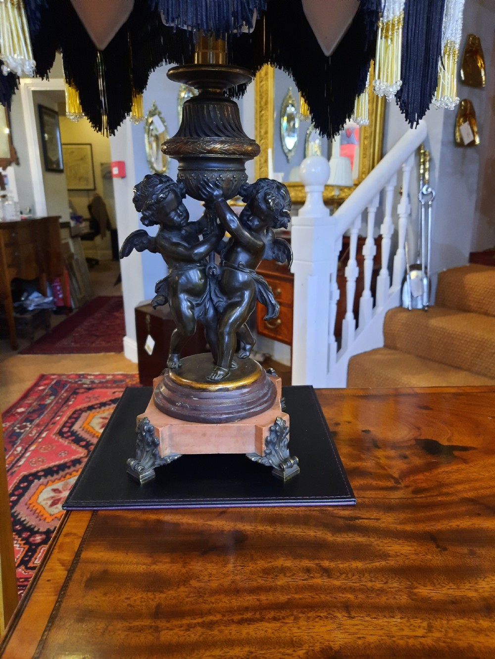 An antique French bronze table lamp with sculpted cherub figures resting on a marble base, topped with a cream and black scalloped fabric shade. View II