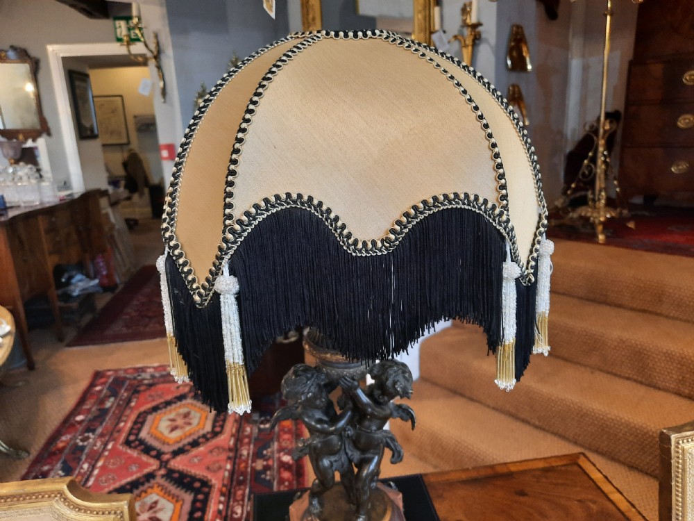 An antique French bronze table lamp with sculpted cherub figures resting on a marble base, topped with a cream and black scalloped fabric shade. View III