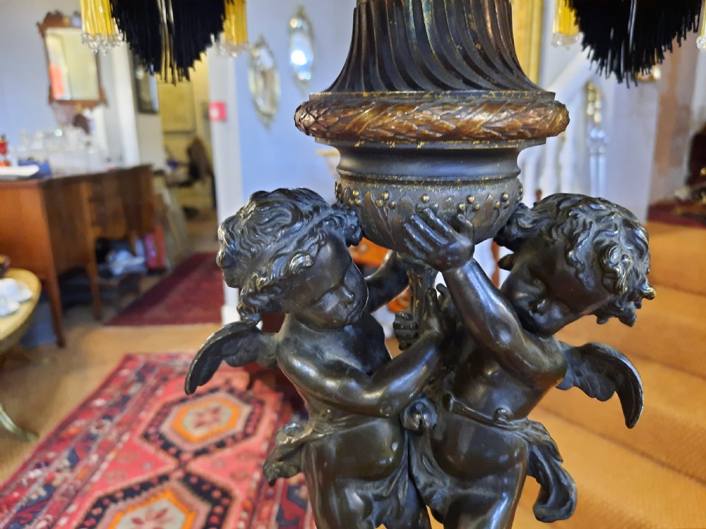 An antique French bronze table lamp with sculpted cherub figures resting on a marble base, topped with a cream and black scalloped fabric shade. View V