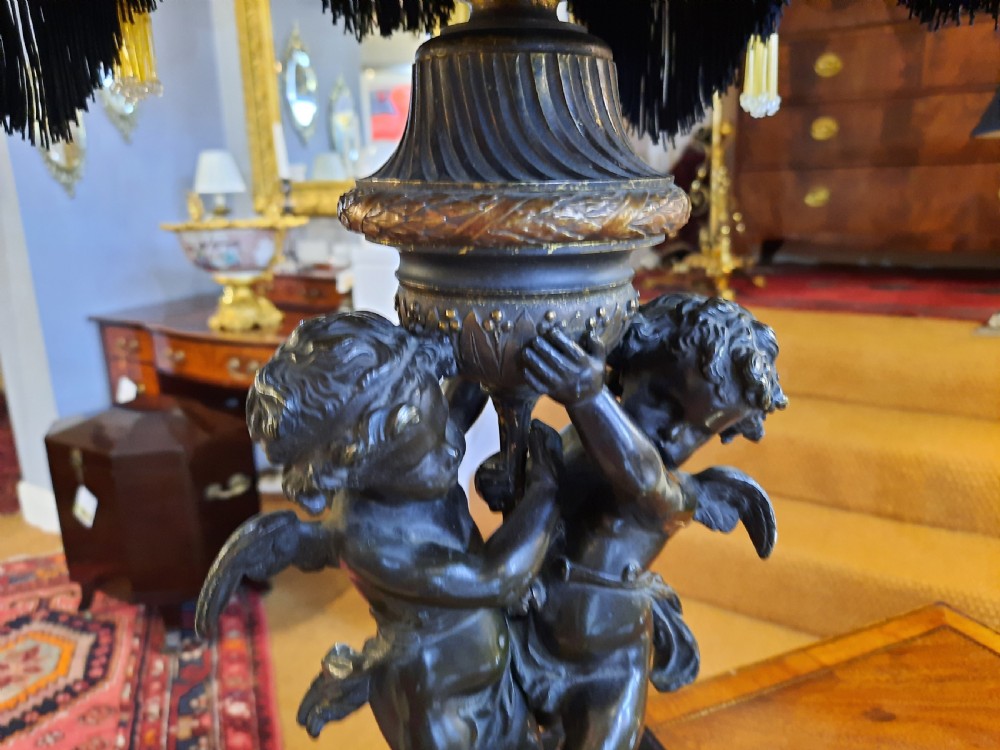 An antique French bronze table lamp with sculpted cherub figures resting on a marble base, topped with a cream and black scalloped fabric shade. View VI