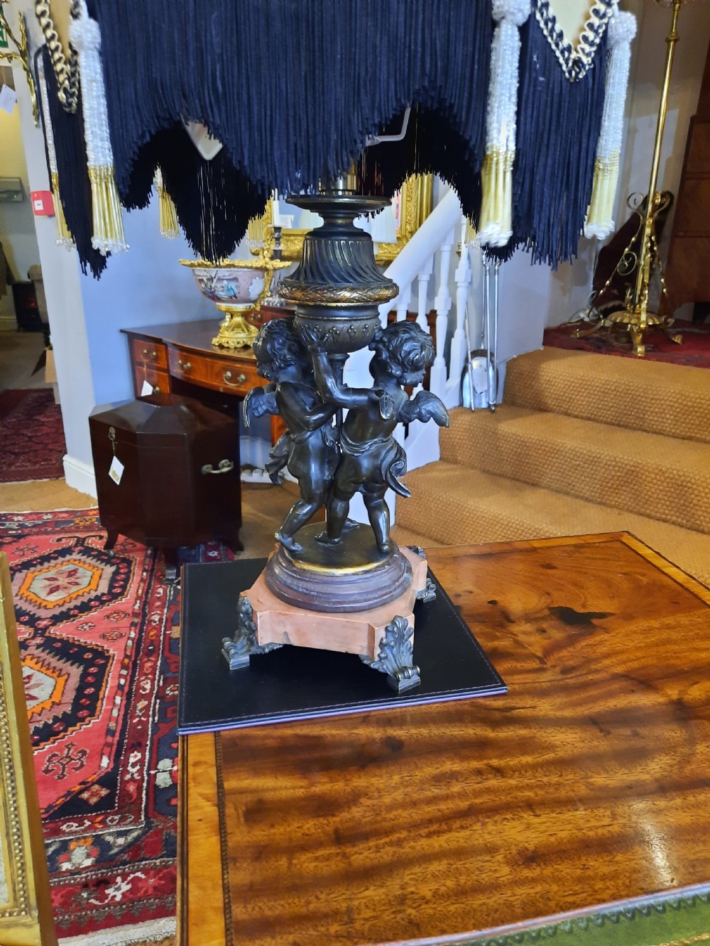 An antique French bronze table lamp with sculpted cherub figures resting on a marble base, topped with a cream and black scalloped fabric shade. View VIII