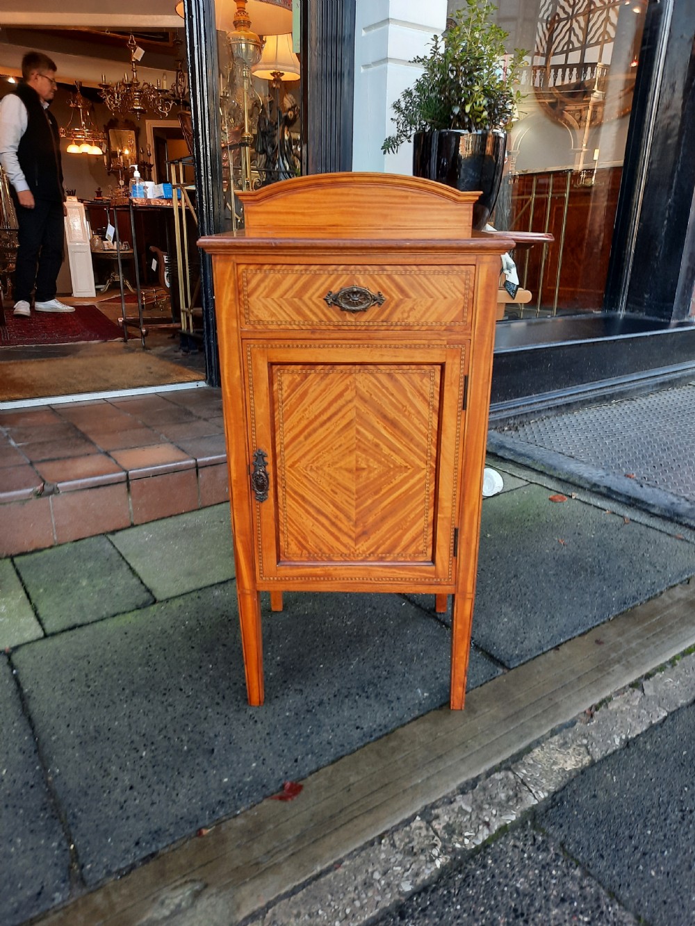 An Edwardian satinwood bedside cabinet featuring chevron pattern veneered door and drawer panels, metal drop handles, and tapered square legs. View II
