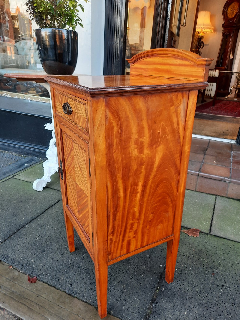 An Edwardian satinwood bedside cabinet featuring chevron pattern veneered door and drawer panels, metal drop handles, and tapered square legs. View III