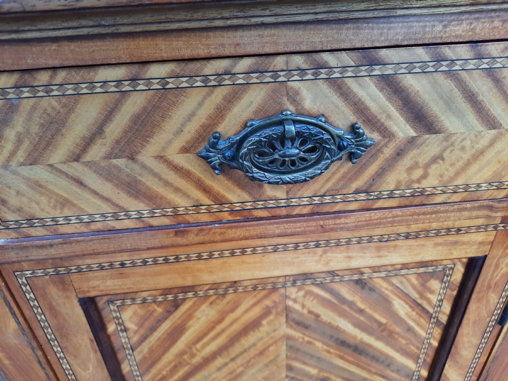 An Edwardian satinwood bedside cabinet featuring chevron pattern veneered door and drawer panels, metal drop handles, and tapered square legs. View V
