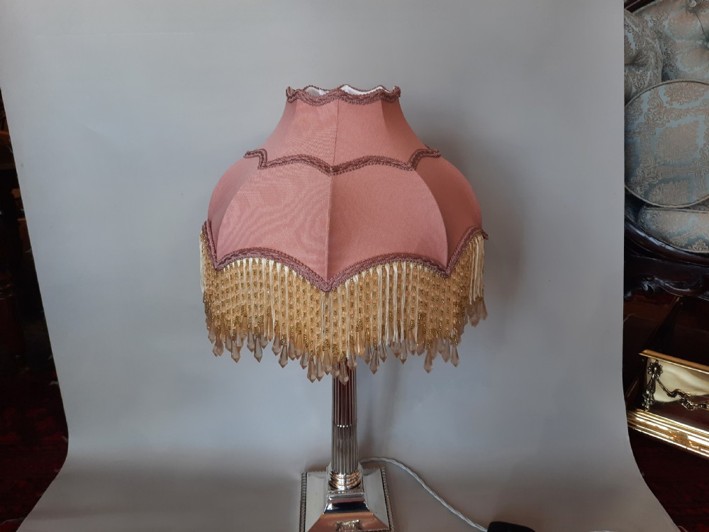 A silver-plated table lamp styled as a fluted Corinthian column with a stepped square base, topped with a scalloped pink fabric shade adorned with long beaded fringe. View VIII