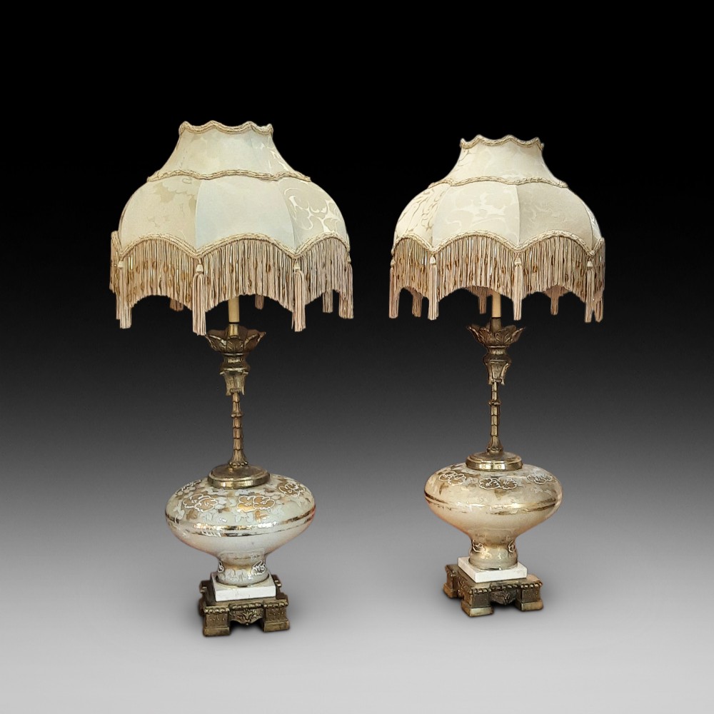 A pair of late Victorian table lamps featuring ornate gilt brass stems, painted glass baluster bases, and cream silk shades with fringe tassels.