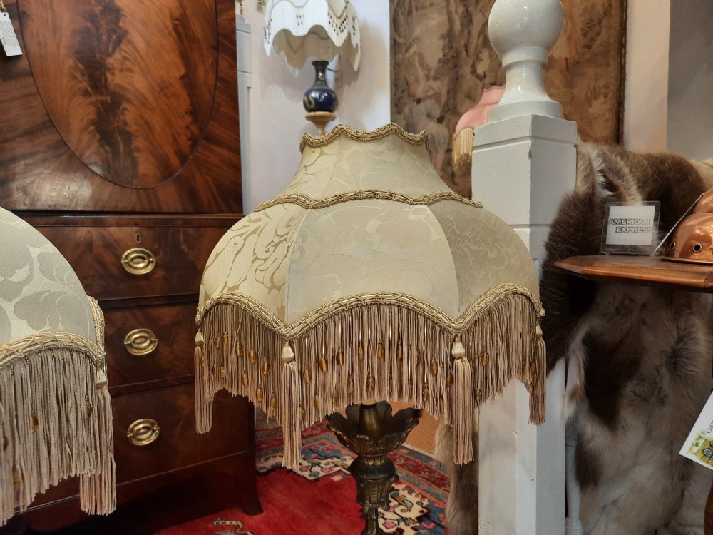 A pair of late Victorian table lamps featuring ornate gilt brass stems, painted glass baluster bases, and cream silk shades with fringe tassels. View V